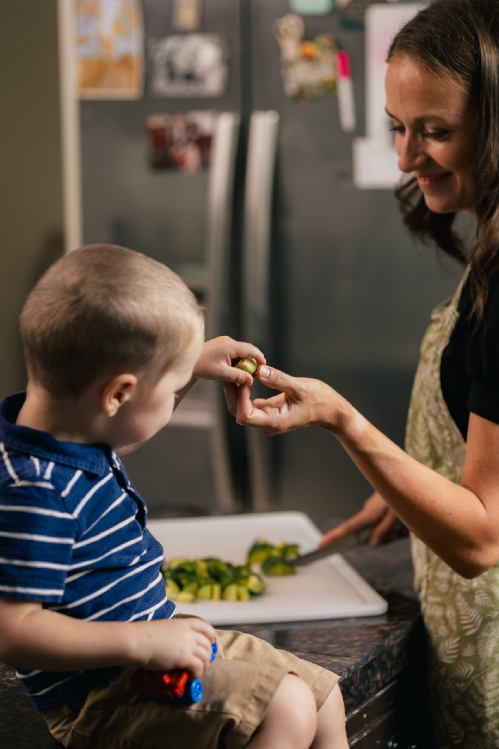 Mother handing a piece of food to her young son in the kitchen while preparing a meal