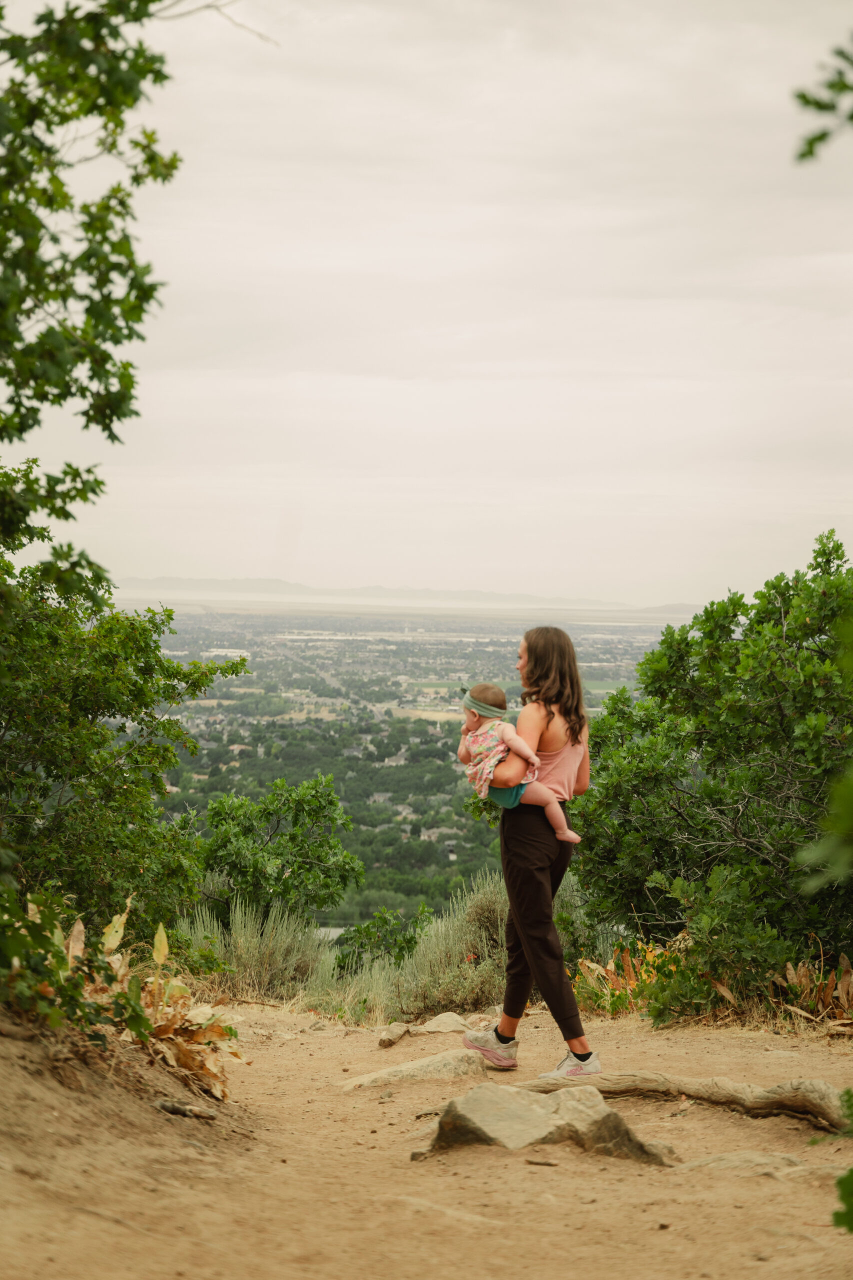 Mother hiking a mountain trail while holding her baby, overlooking a valley view, representing general wellness